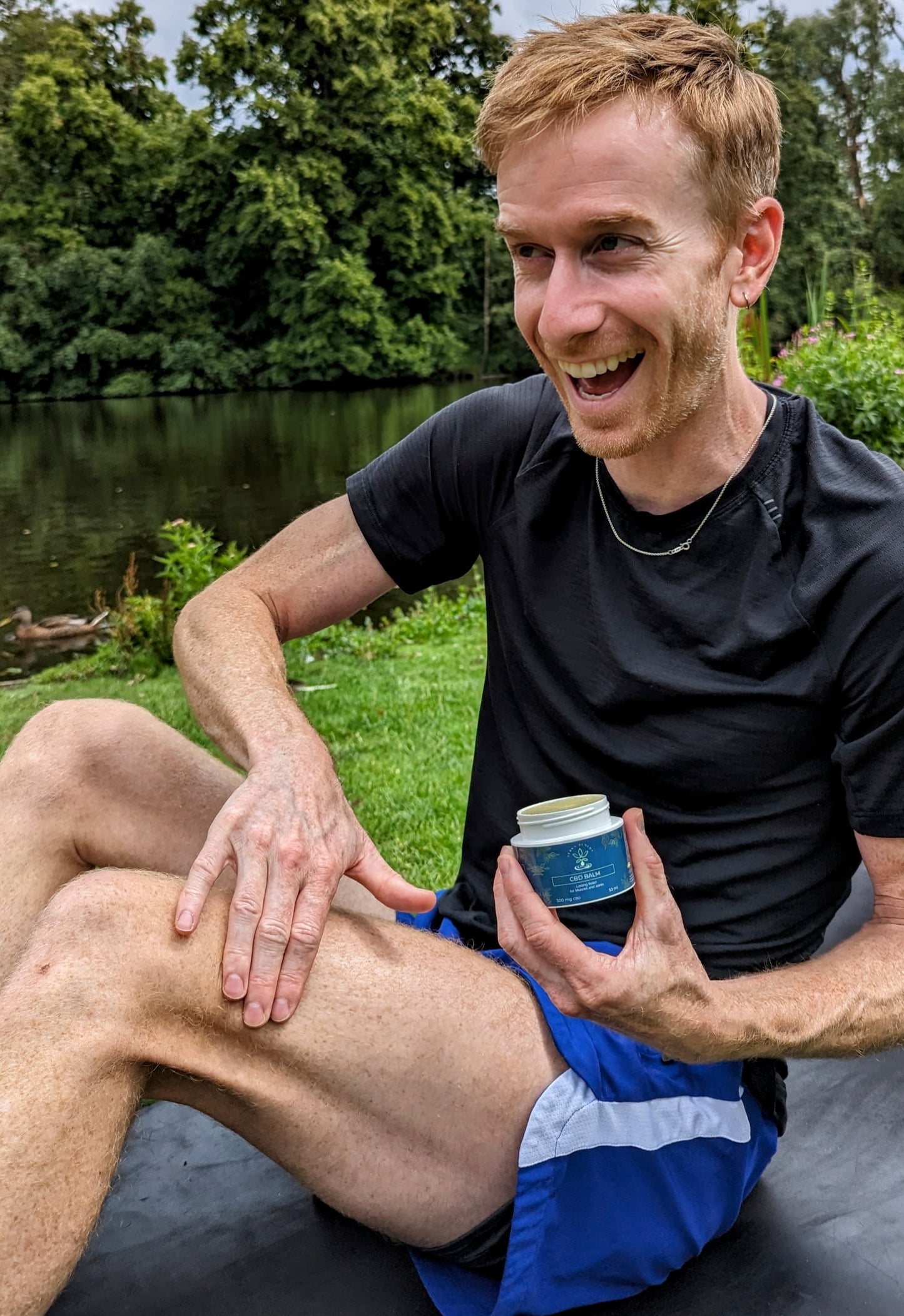 
                  
                    An athlete sitting outdoors by a pond, smiling while applying CBD balm to his leg. He holds a jar of the balm, showcasing its use for muscle relief and recovery after physical activity.
                  
                