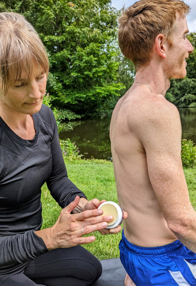 
                  
                    A person applying CBD balm to another individual’s back outdoors by a pond. The balm is used for targeted muscle relief, showcasing its use for soothing sore muscles and reducing discomfort naturally.
                  
                